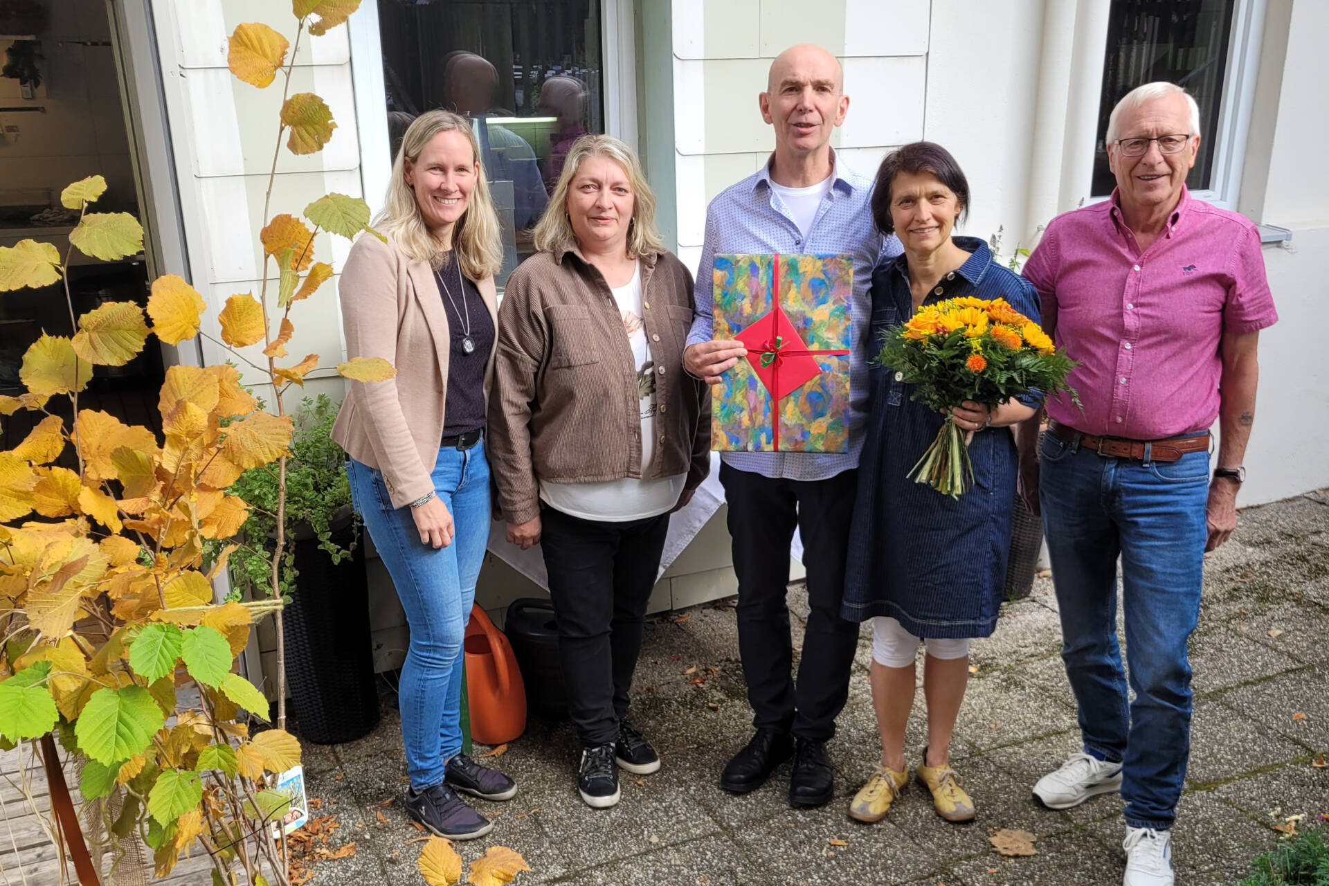 Fünf Personen stehen vor einem Haus, eine hält ein Geschenk und eine einen Blumenstrauß.